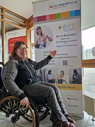 EUTB-Beraterin im Rollstuhl zeigt auf ein Poster. Dort steht; Bist du Behindert? Macht nix! Fürs Studium reichts! Tipps und Tricks bei uns!